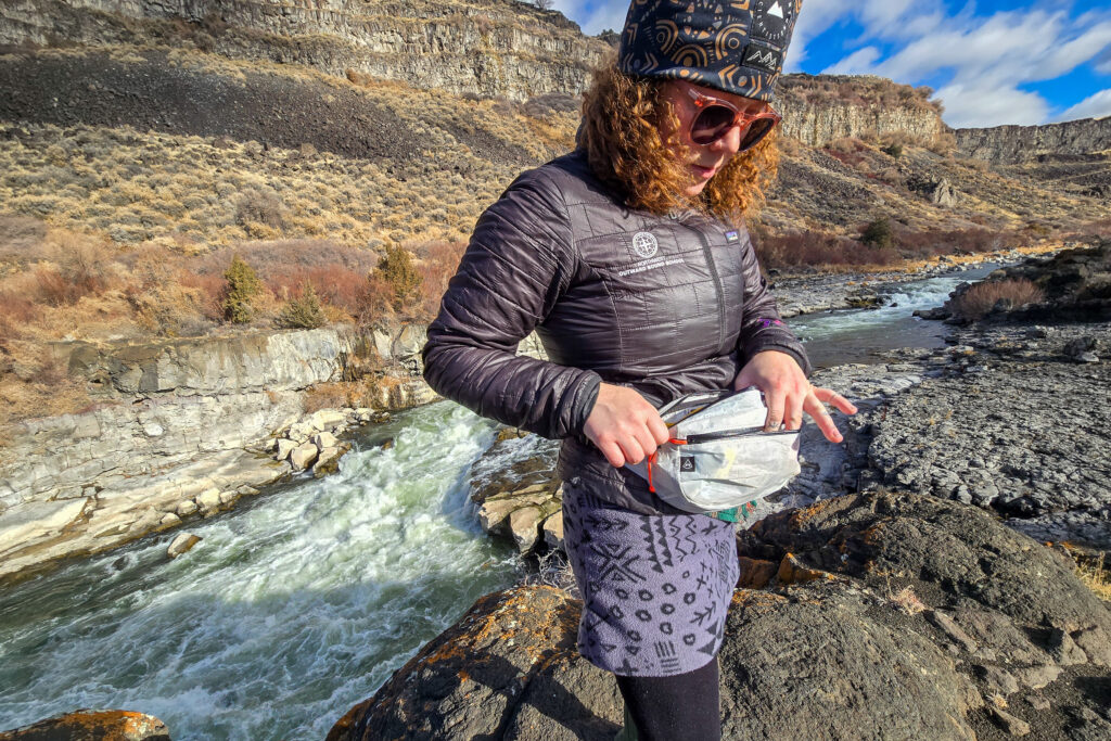 woman unzipping the hyperlite mountain gear versa fany pack next to a rushing white water river