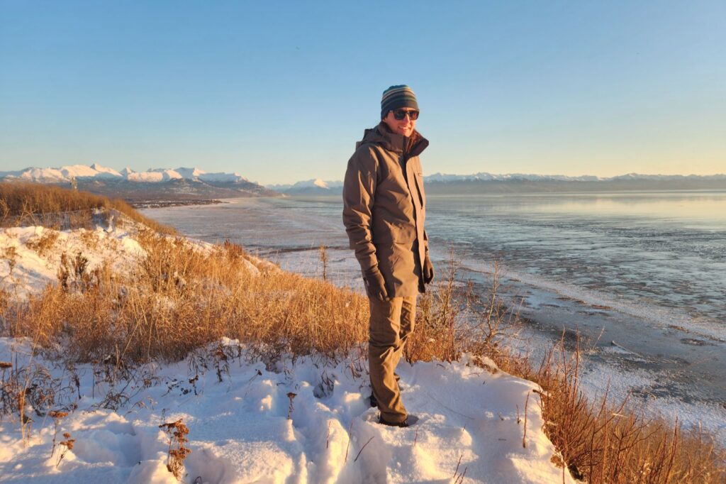 A man stands on a snowy hill overlooking the ocean at sunset.