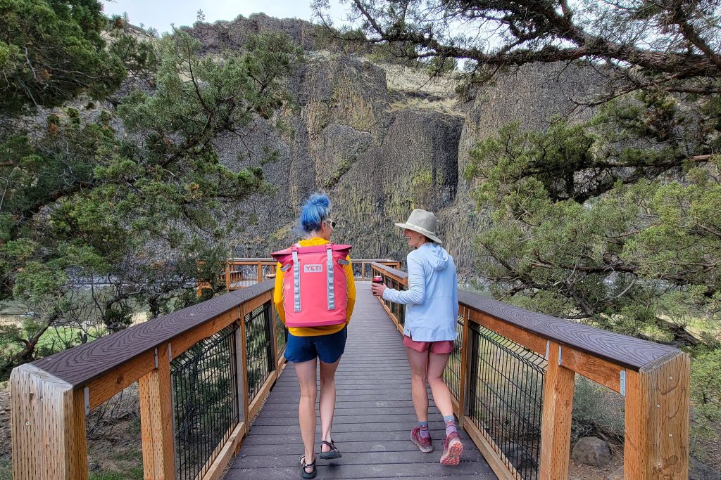 two women walk across a bridge wearing a cooler bag and holding a drink