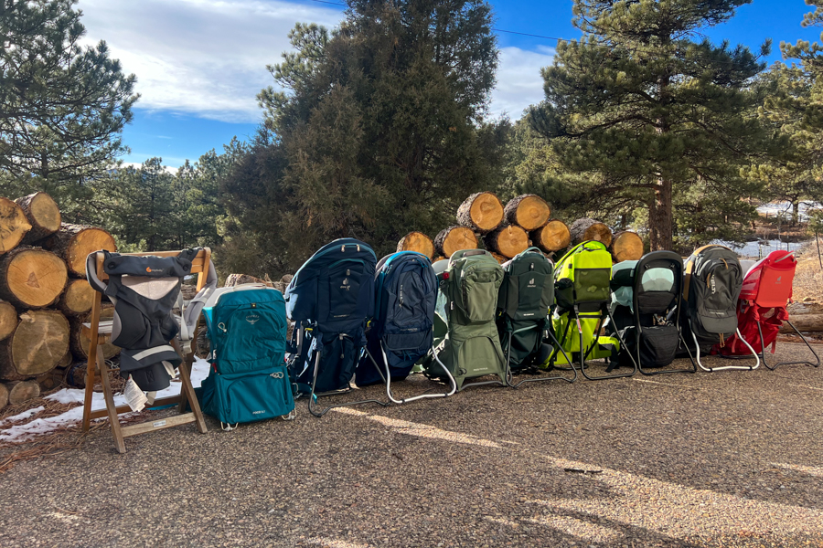 10 baby backpack carriers lineup up in front of a wood pile with pine trees in the background.