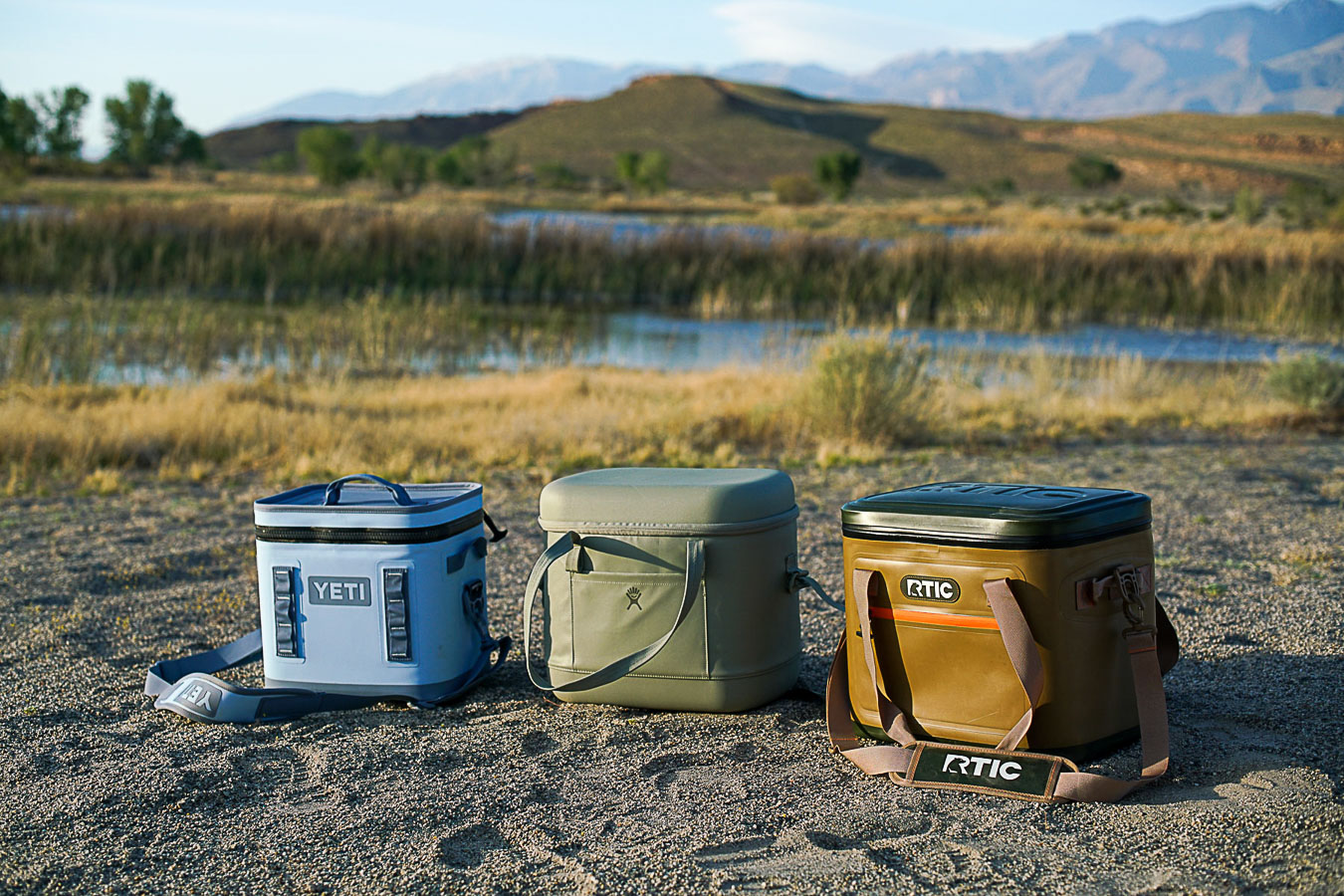 The YETI Hopper Flip 12, Hydro Flask Carry Out 20L, and RTIC Soft Pack cooler sit on the ground with a body of water and mountains in the background.