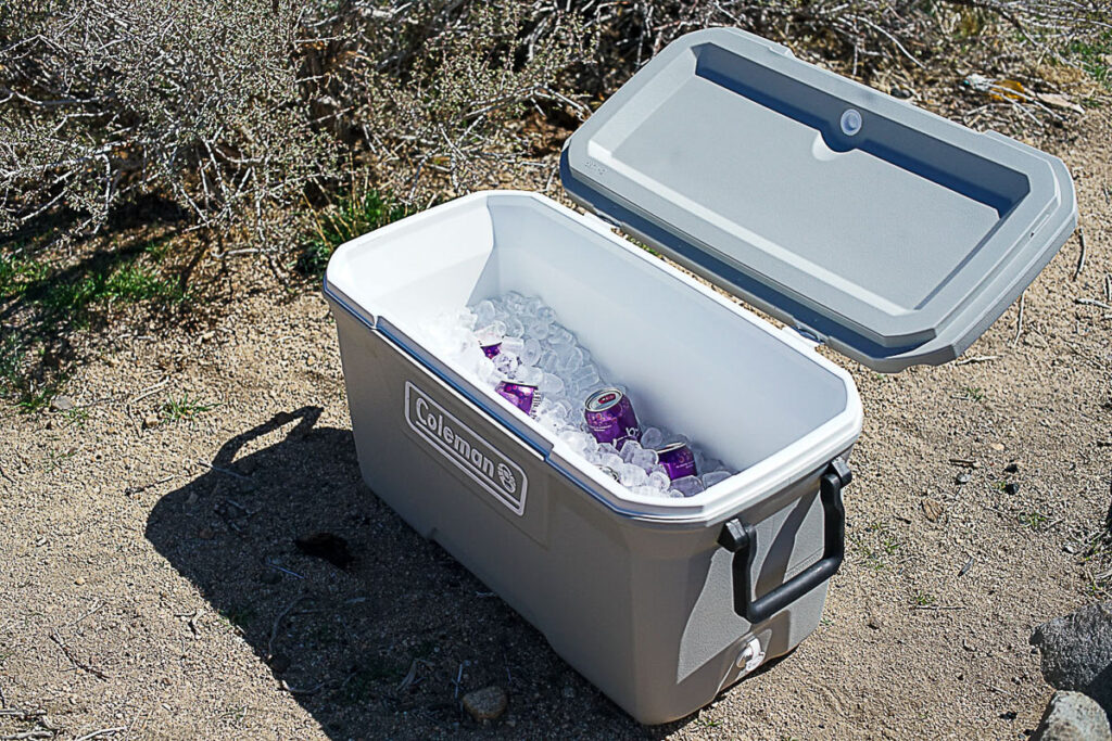 The Coleman 316 Series 70QT cooler sits on the ground with its lid open. The cooler is full of ice and some canned drinks are visible.