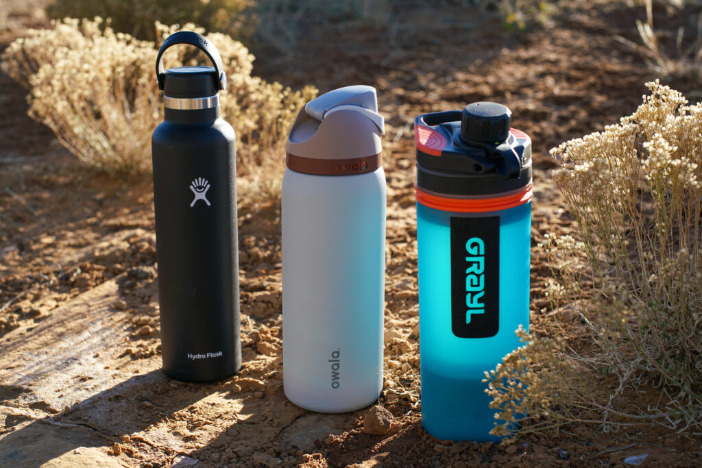 The Hydro Flask Standard Mouth, Owala FreeSip Vacuum Insulated, and Grayl GeoPress sit on the ground with bushes behind them.