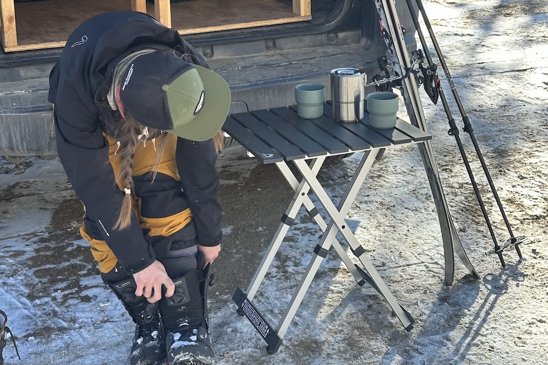 A person bends down, sitting on the tailgate of a truck, taking off their boots. The table has a stainless steel mug and two teal cups on it. Skis and poles are on the right of the table.