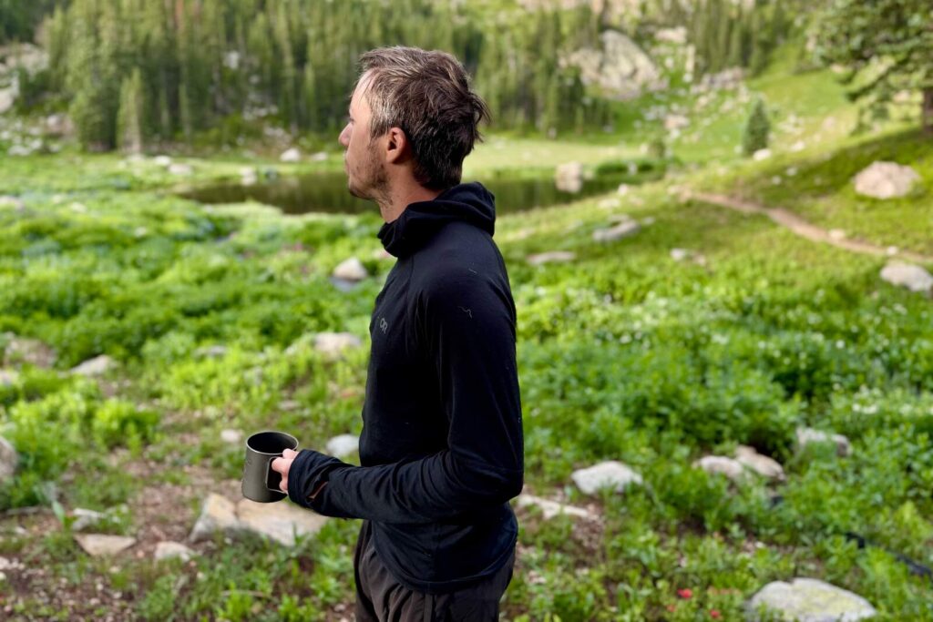 a hiker in a hooded black base layer stands with a coffee mug in an alpine environment