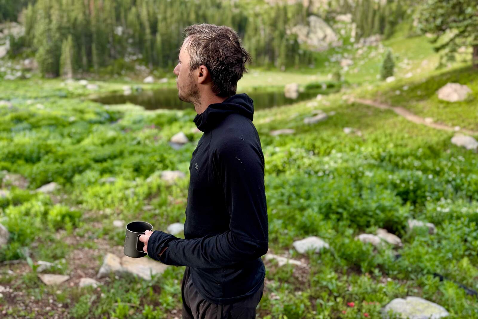 a hiker in a hooded black base layer stands with a coffee mug in an alpine environment
