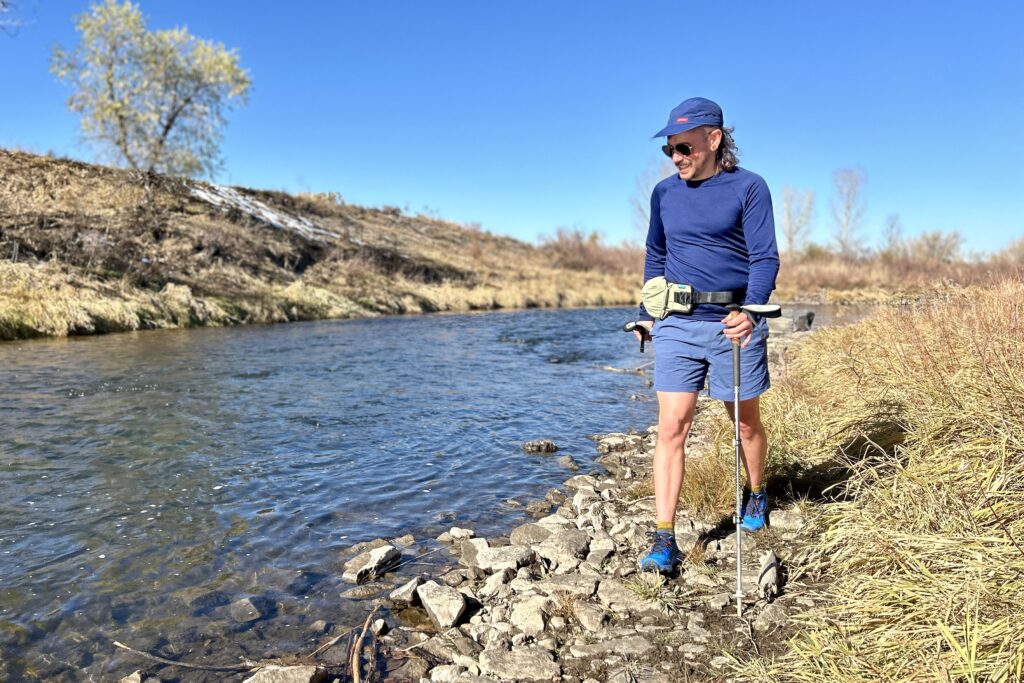A man hikes along a river in a wilderness area in shorts and he is wearing all blue.