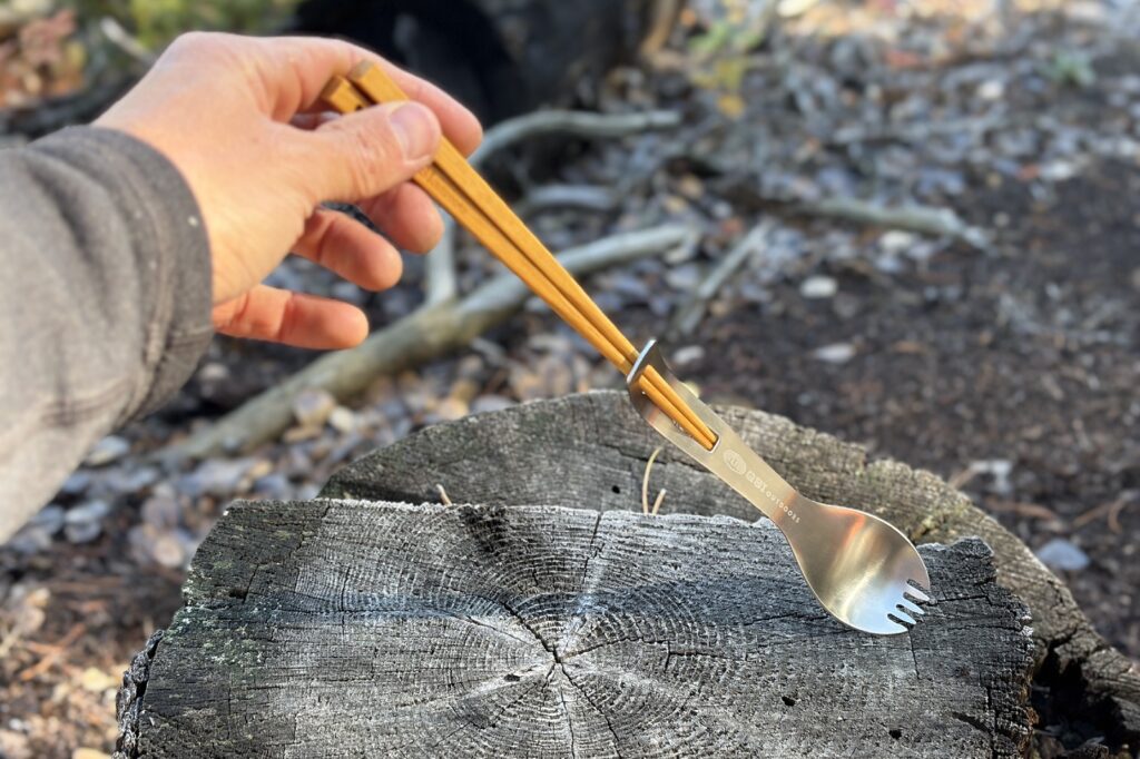 a hand holds a chopstick spork combo on a tree stump