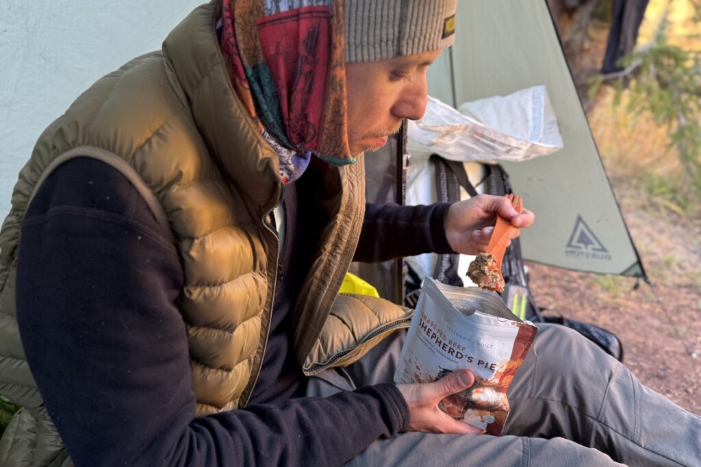 a hiker sits in a tent and eats a backpacking meal with a long plastic spork