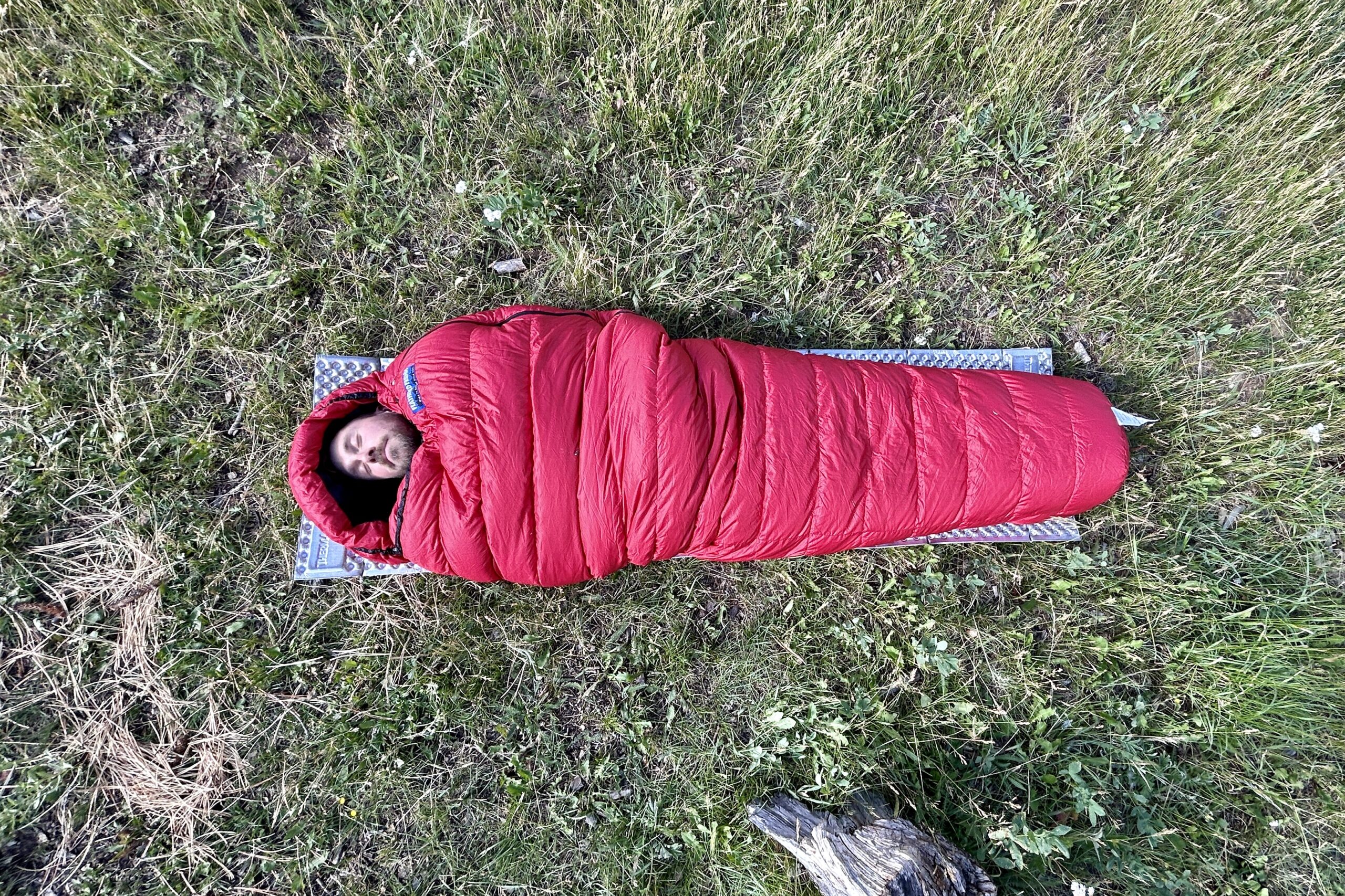 An mage from above of a man resting in a sleeping bag on a foam pad with the hood up. He is cowboy camping on the grass in the wilderness.