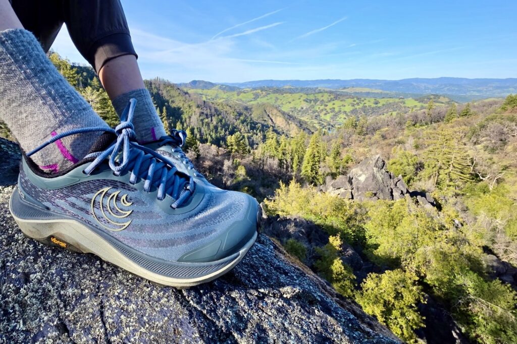 A person sitting on an overlook with a beautiful scene in the background, with a focus on her hiking shoes.