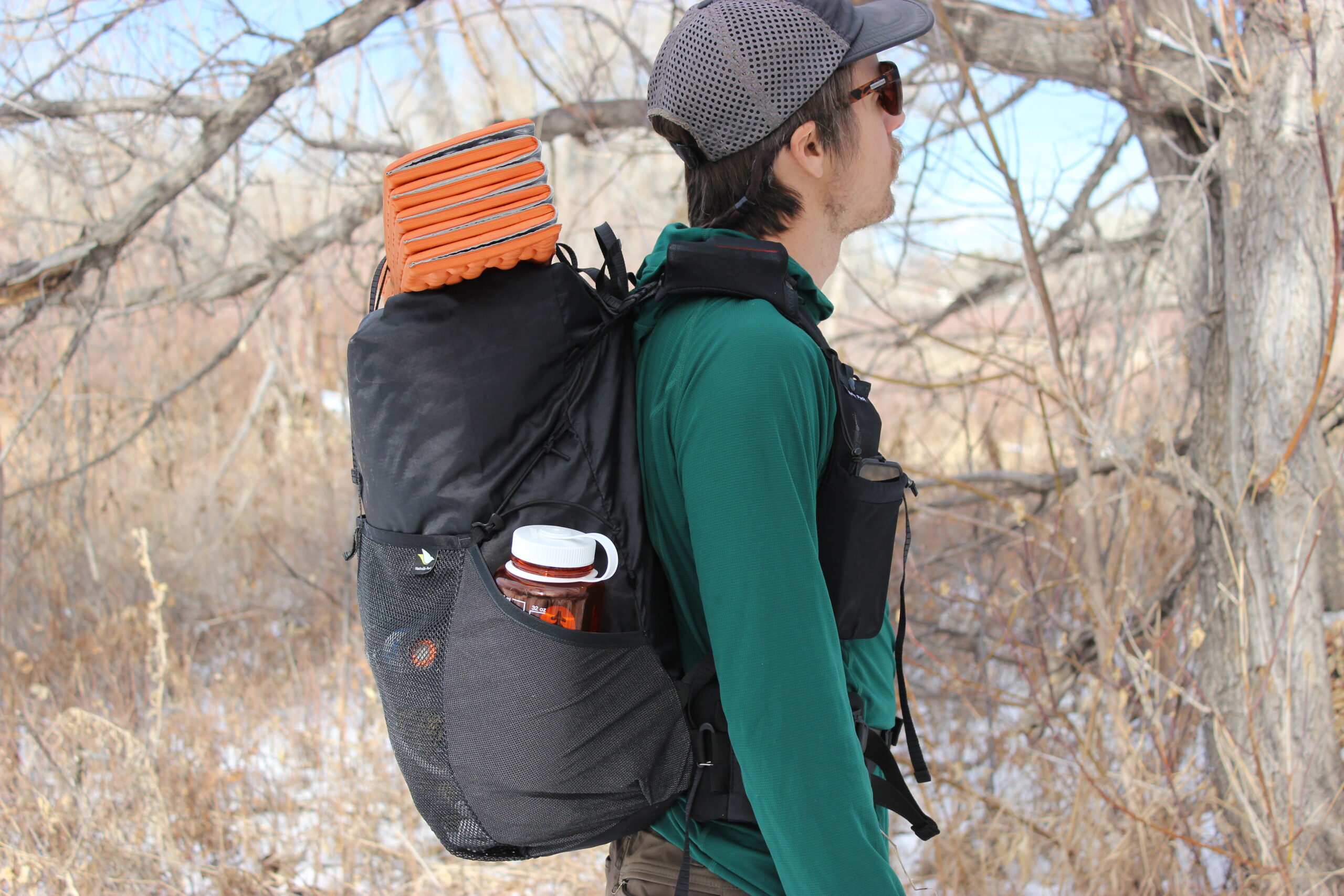 A profile shot of a hiker wearing the Nashville Pack Cutaway with an orange sleeping pad on top and an orange water bottle in the side pocket.