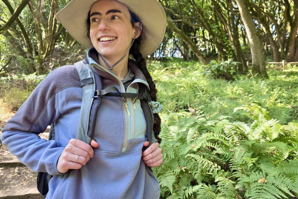 Woman stands on a trail wearing a backpack over her fleece jacket.