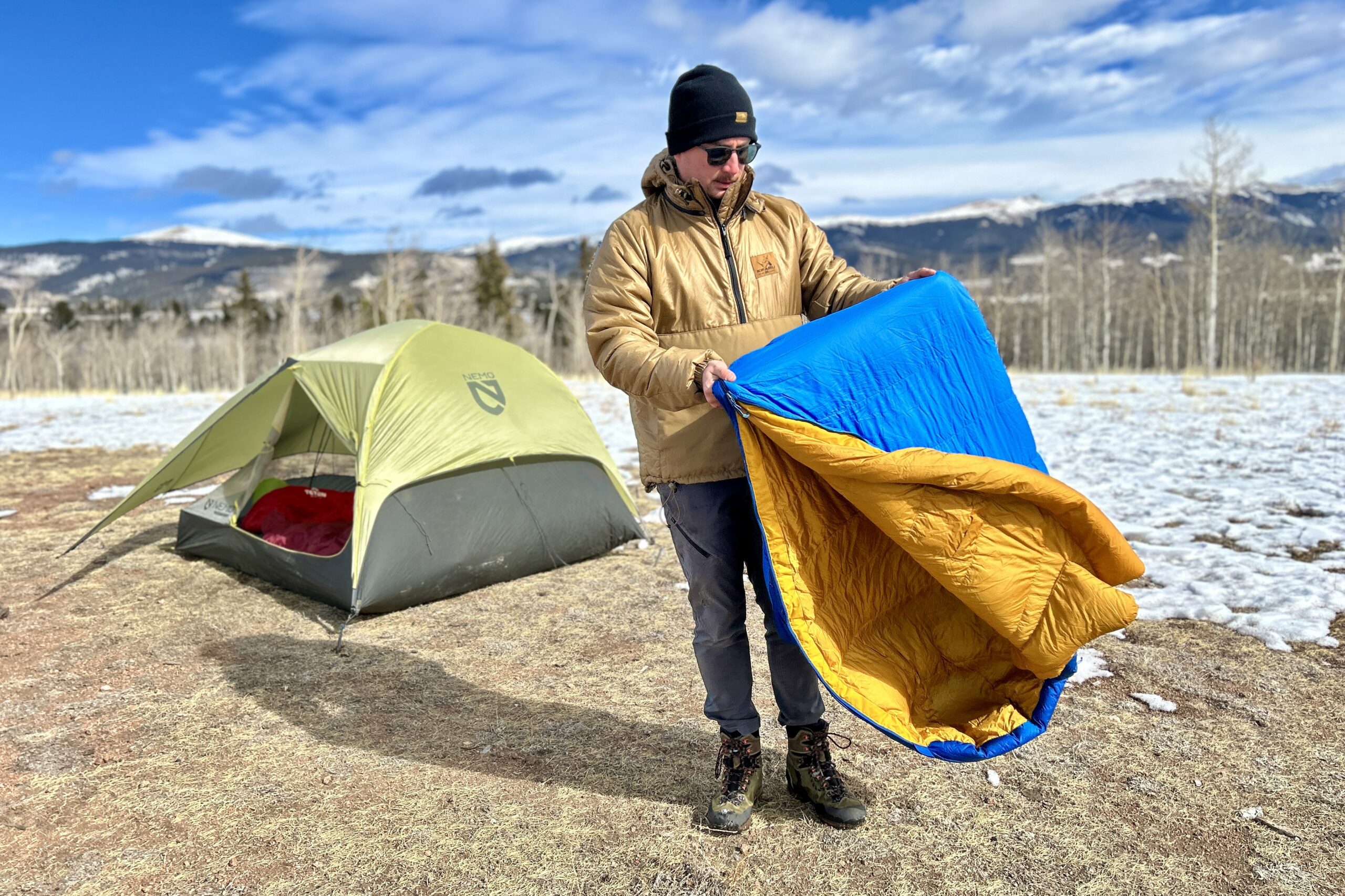 A man fluffs a sleeping bag with a camping tent behind him in a snowy mountain landscape.