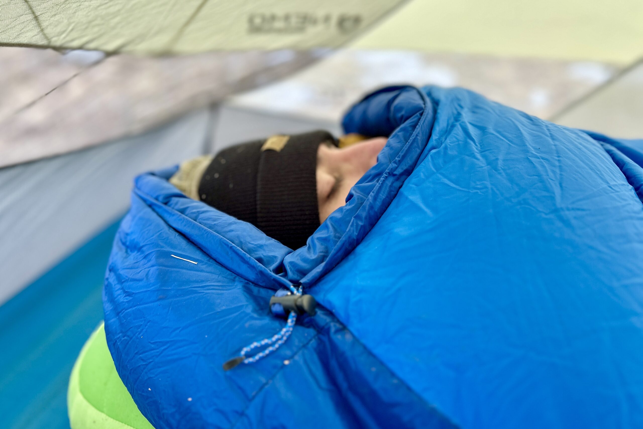 Close-up profile image of a person's face barely peeking out of a closed mummy bag with the mummy hood up and the bag closed to the top.