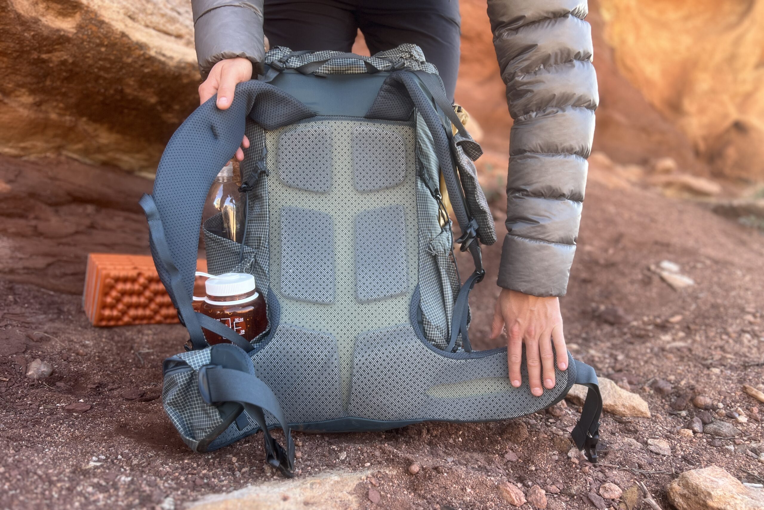 A hiker's hands show the supportive back panel of a backpack with red rocks in the background.