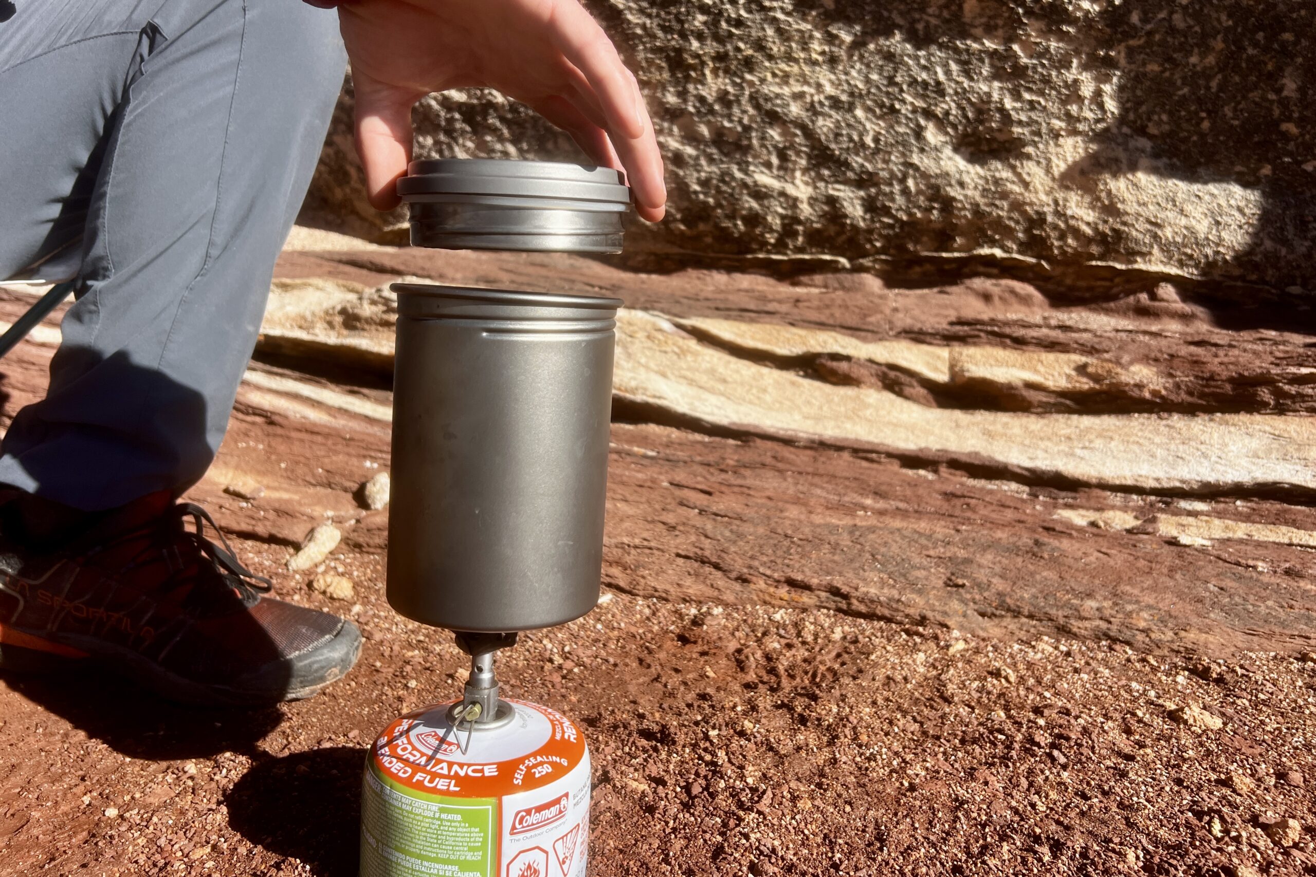A hand pulls the screw-on lid off a backpacking pot against a red rocks background.