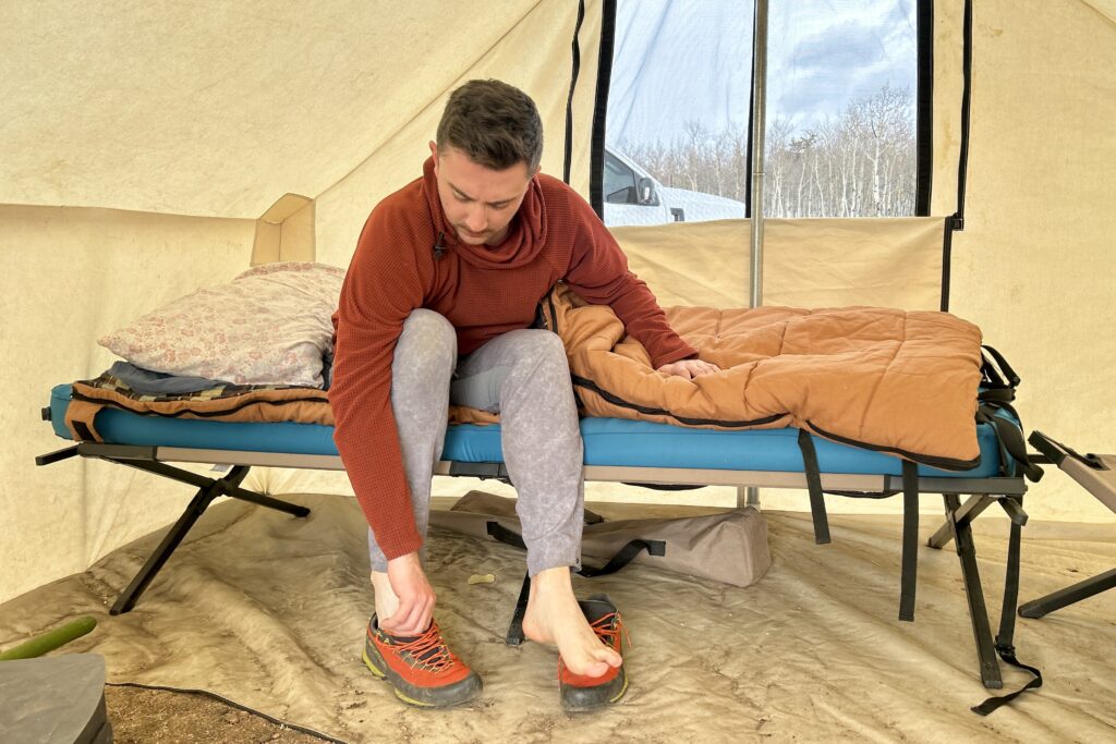 A man sits on a cot with a pad and sleeping bag on it inside of a canvas tent while putting on his shoes.