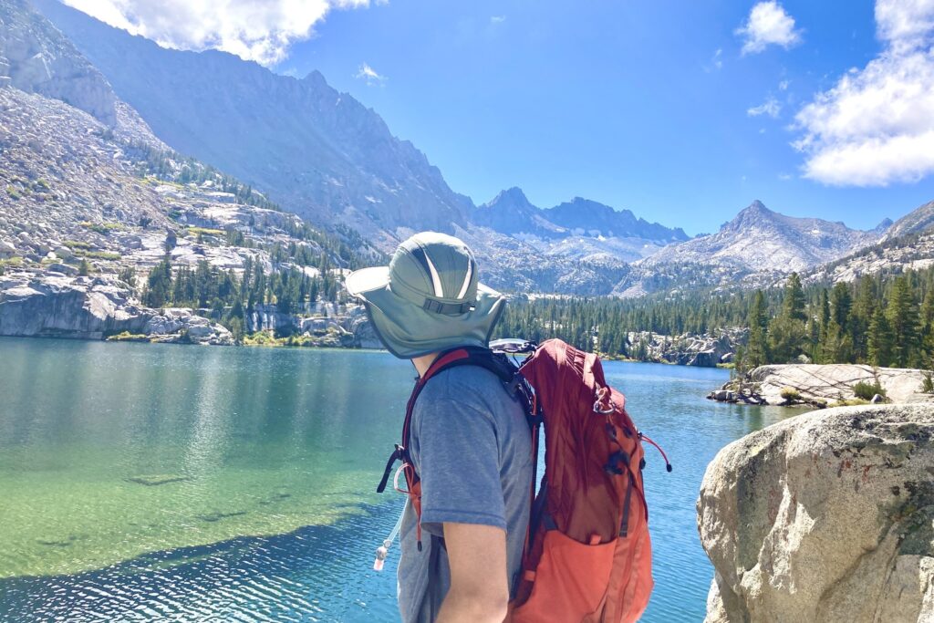 A man stands looking out an alpine lake with mountains in the background.