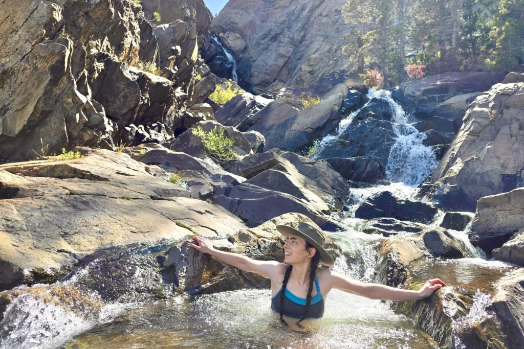 A woman sits in a pool of water below a waterfall wearing her Tilley hat.
