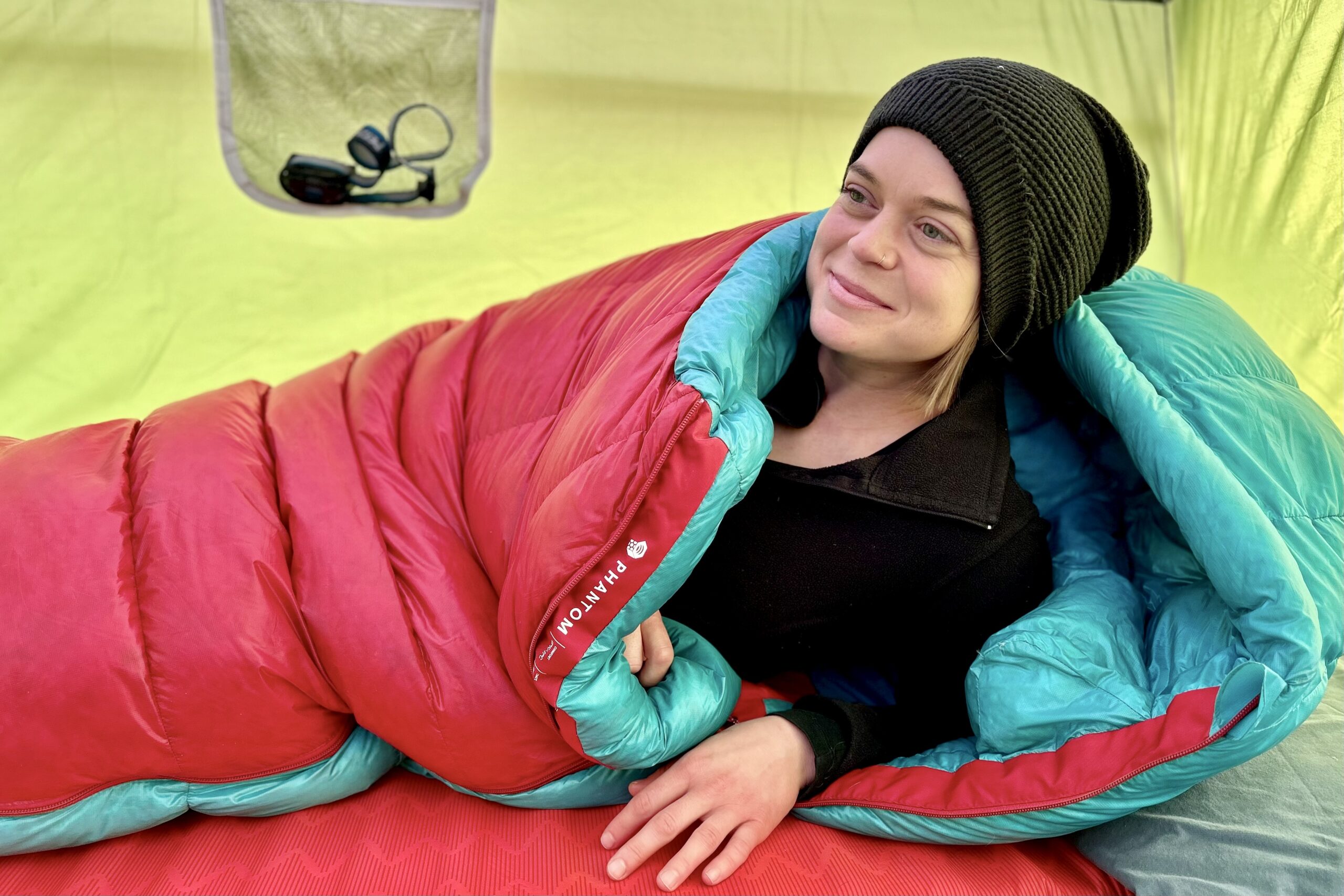 A woman in a sleeping bag sits up on one arm and looks into the distance.