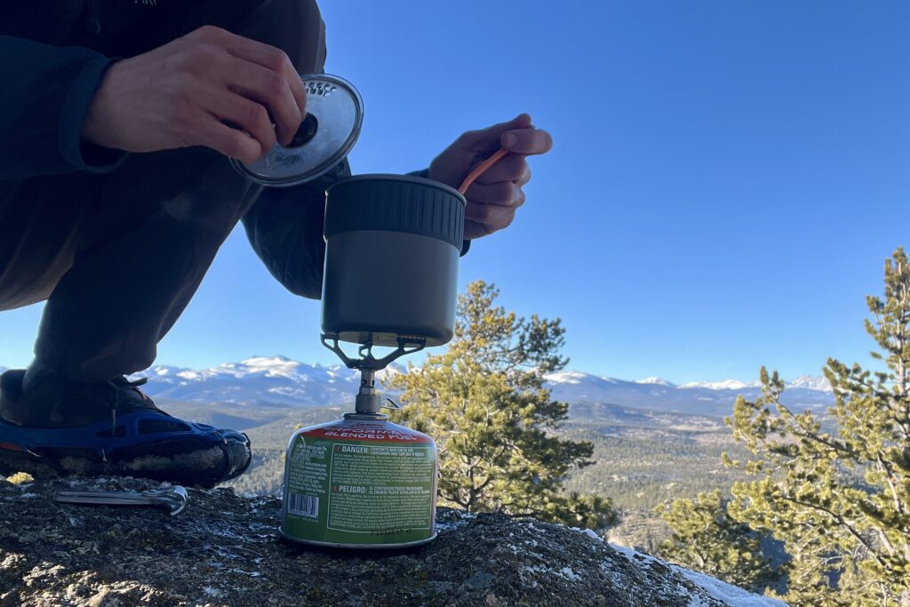 A hiker crouches and cooks with the trail mini solo on a backpacking stove with a mountainous background.