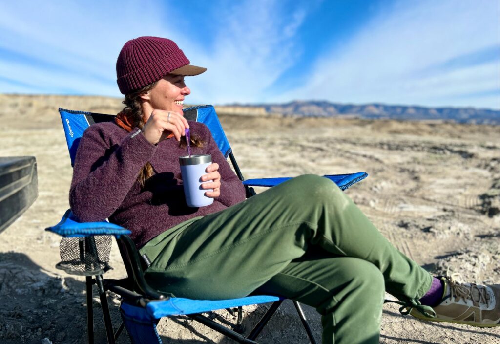 A woman in a camp chair in a desert setting holds a mug and smiles while looking off into the distance