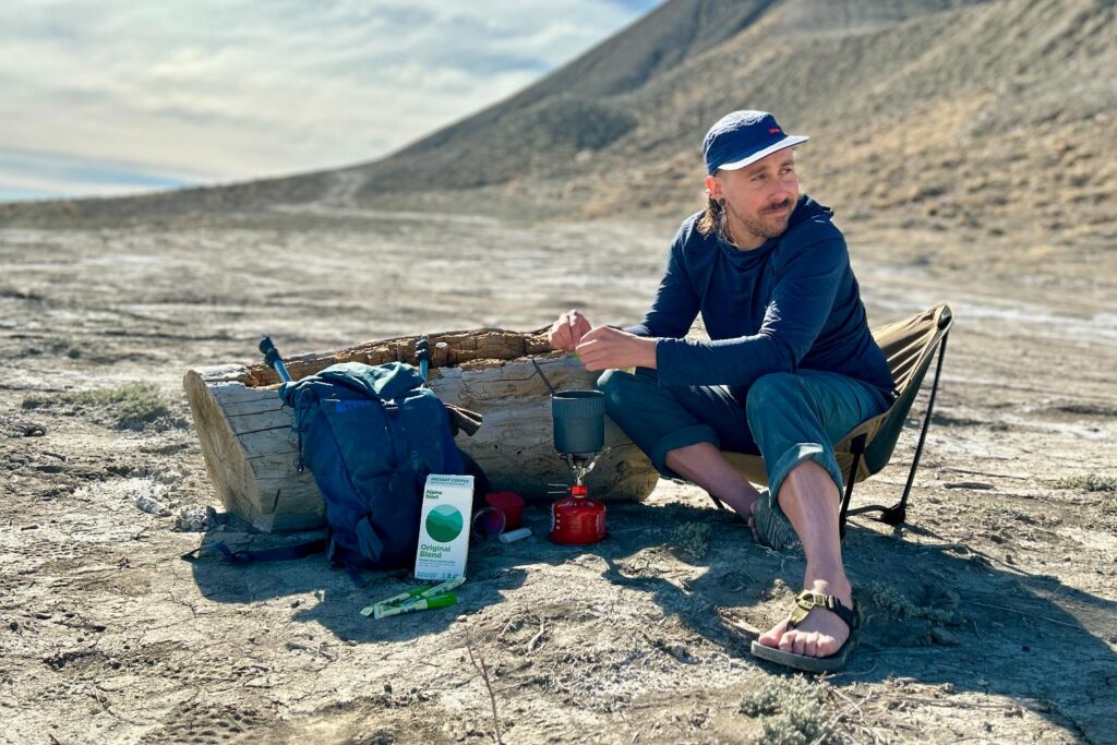 A man sits in a backpacking chair and heats up a pot of water to make coffee with his backpack next to him.
