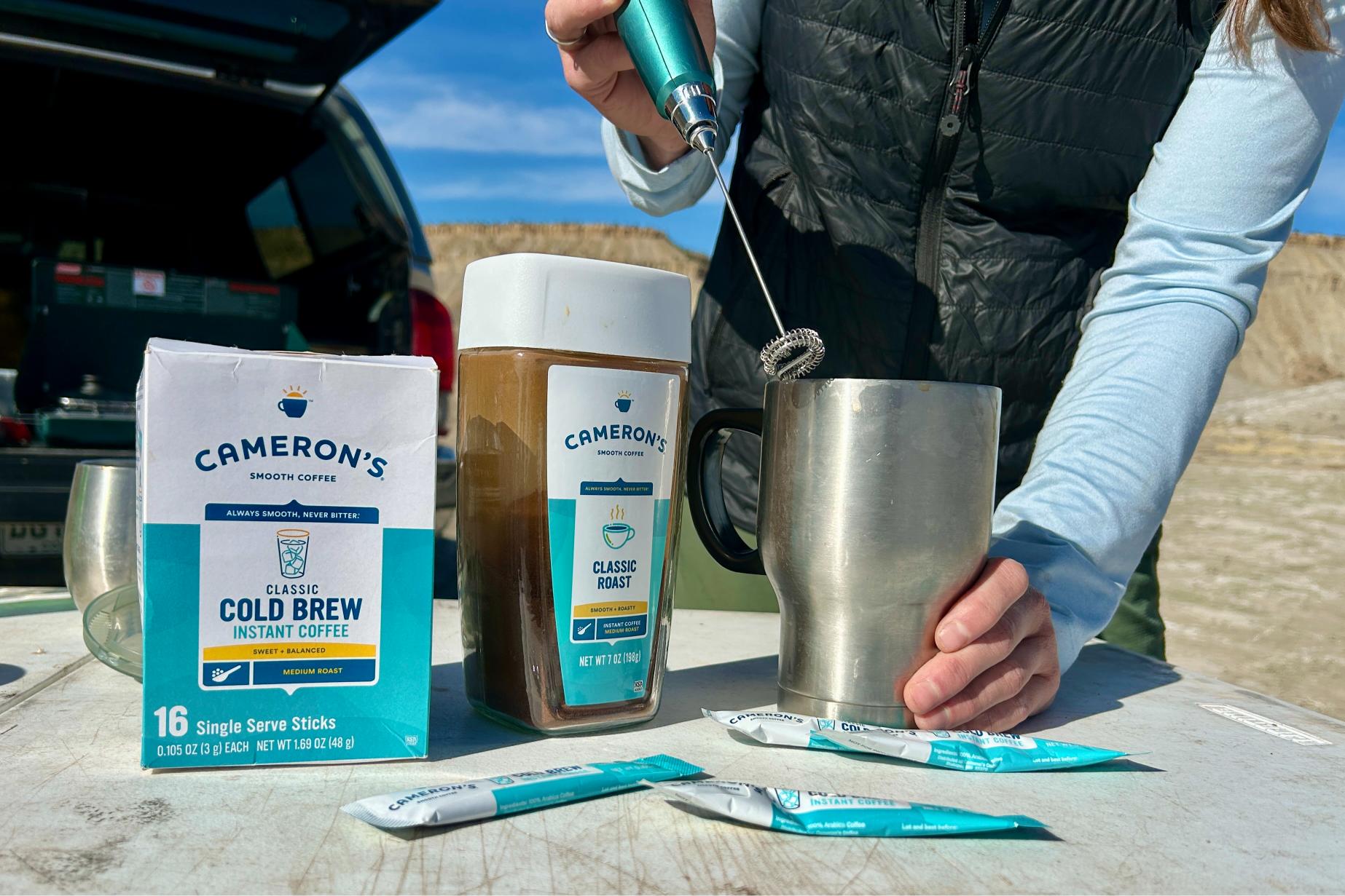 Up-close image of a person using a frother to mix up a cup of instant coffee with bulk and packets of Camerons's next to them on a table