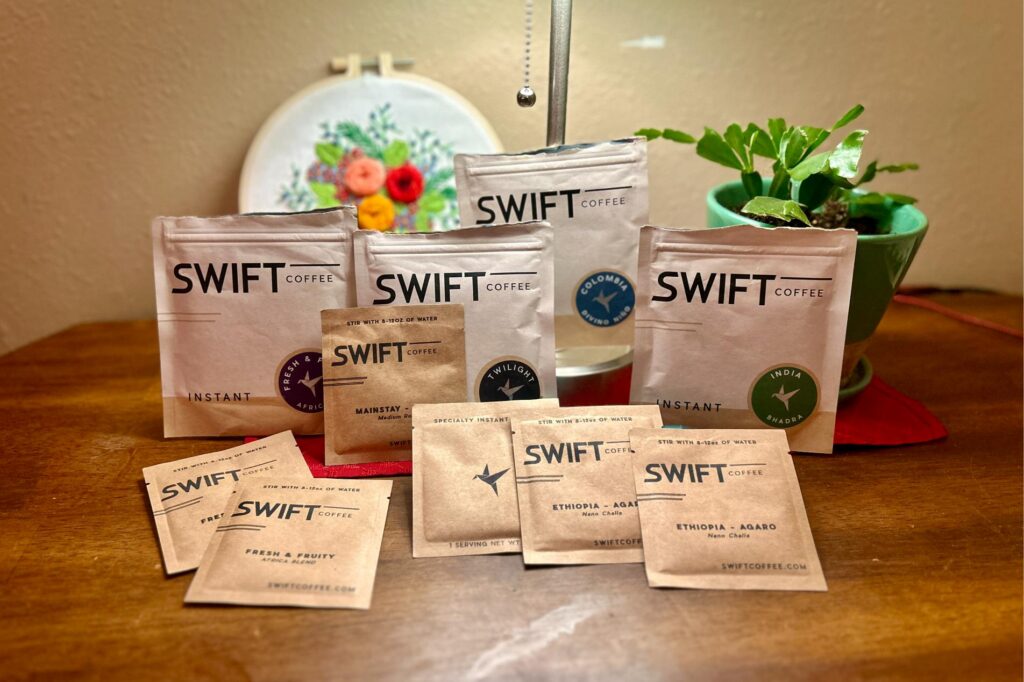 Different varieties of Swift instant coffee staged in front of a plant and a lamp on a table.