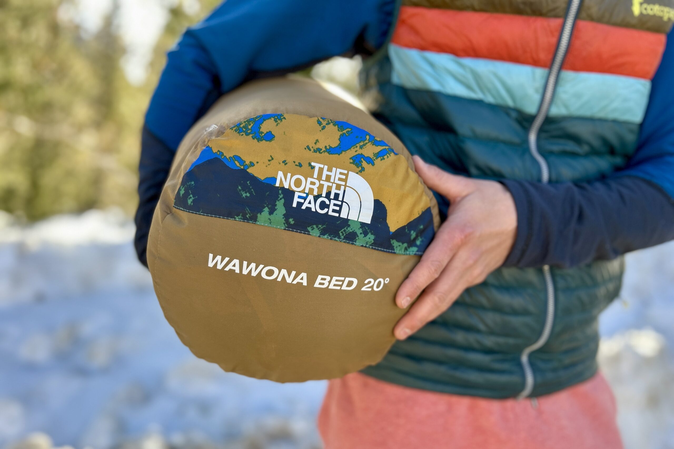 A person holds a sleeping bag in a stuff sack under their arm so the name and logo are visible.