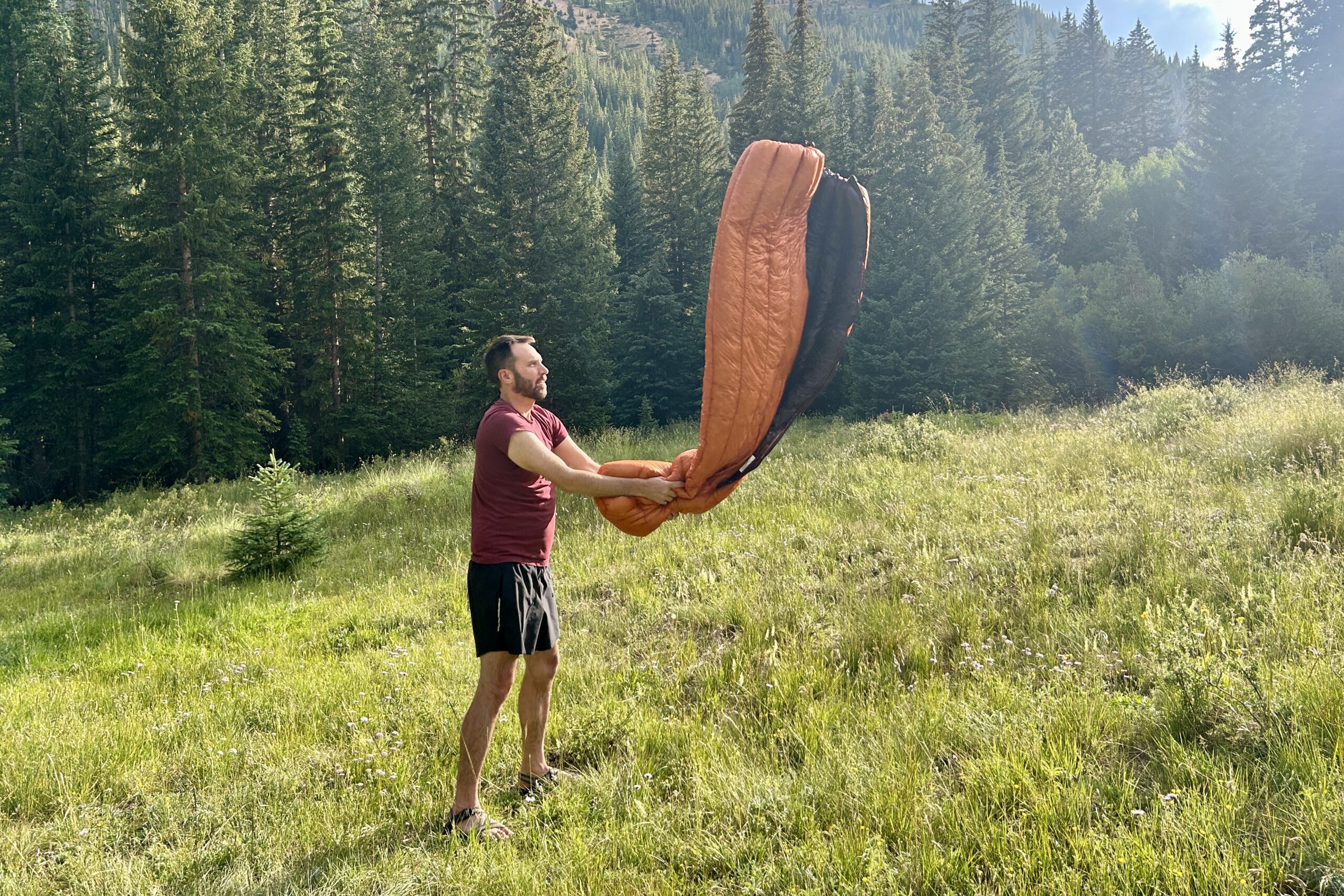 A man shakes out a sleeping bag in a grassy field on a hot summer day.