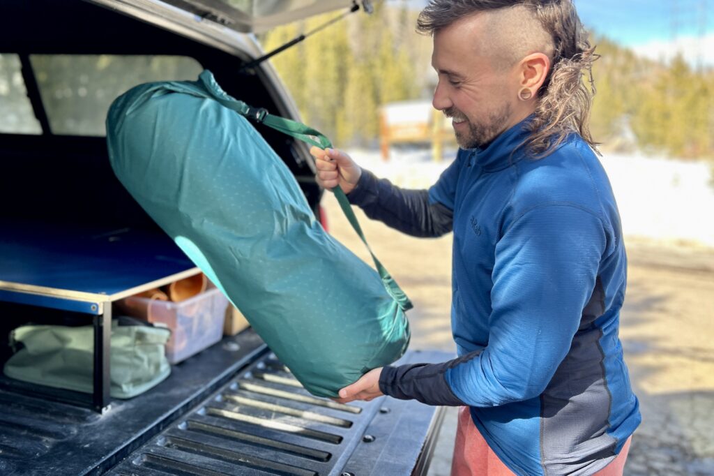 A man smiles as he sets the large storage duffel of a camping mattress in the back of a truck.