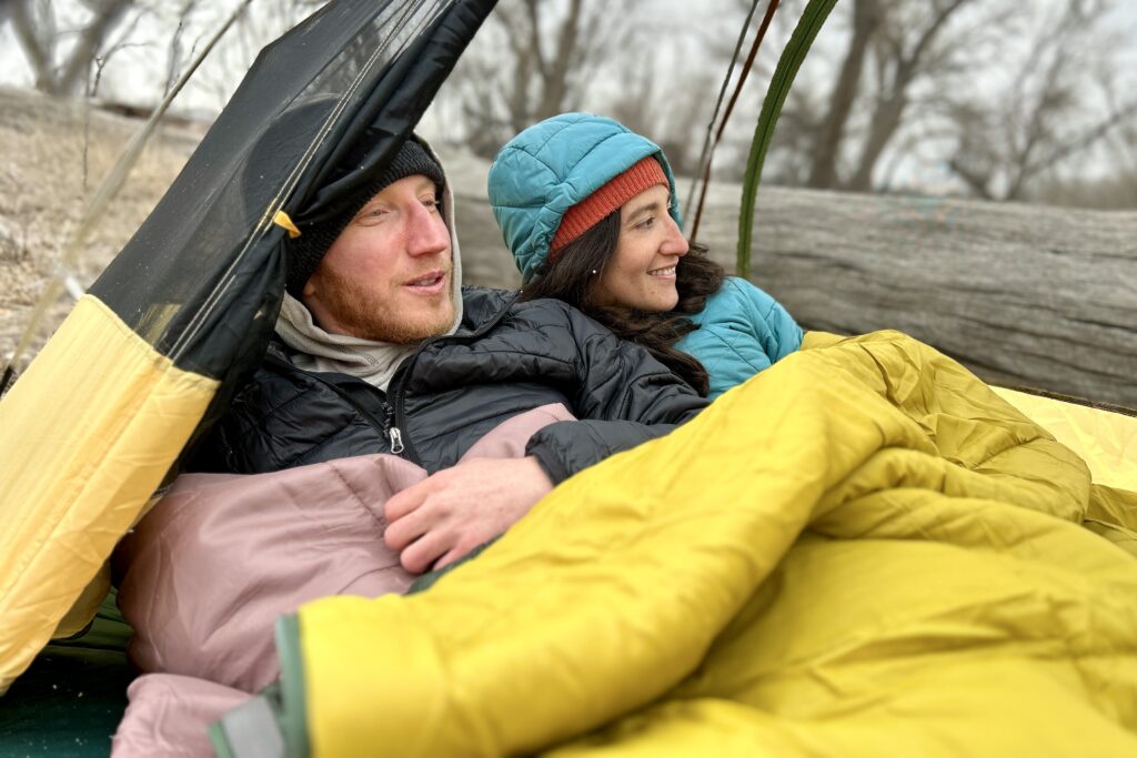 Close-up image of two people in a double sleeping bag in a tent smiling and looking off-camera.
