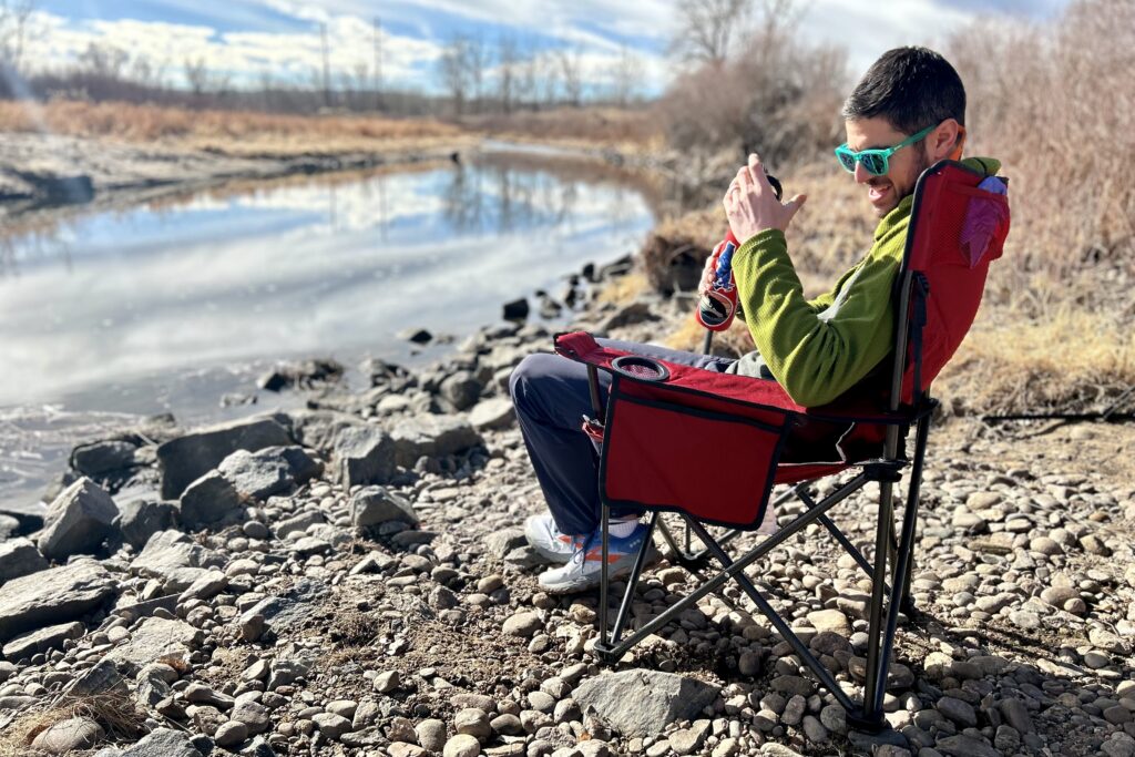 A man laughs while sitting in a camping chair next to a river.
