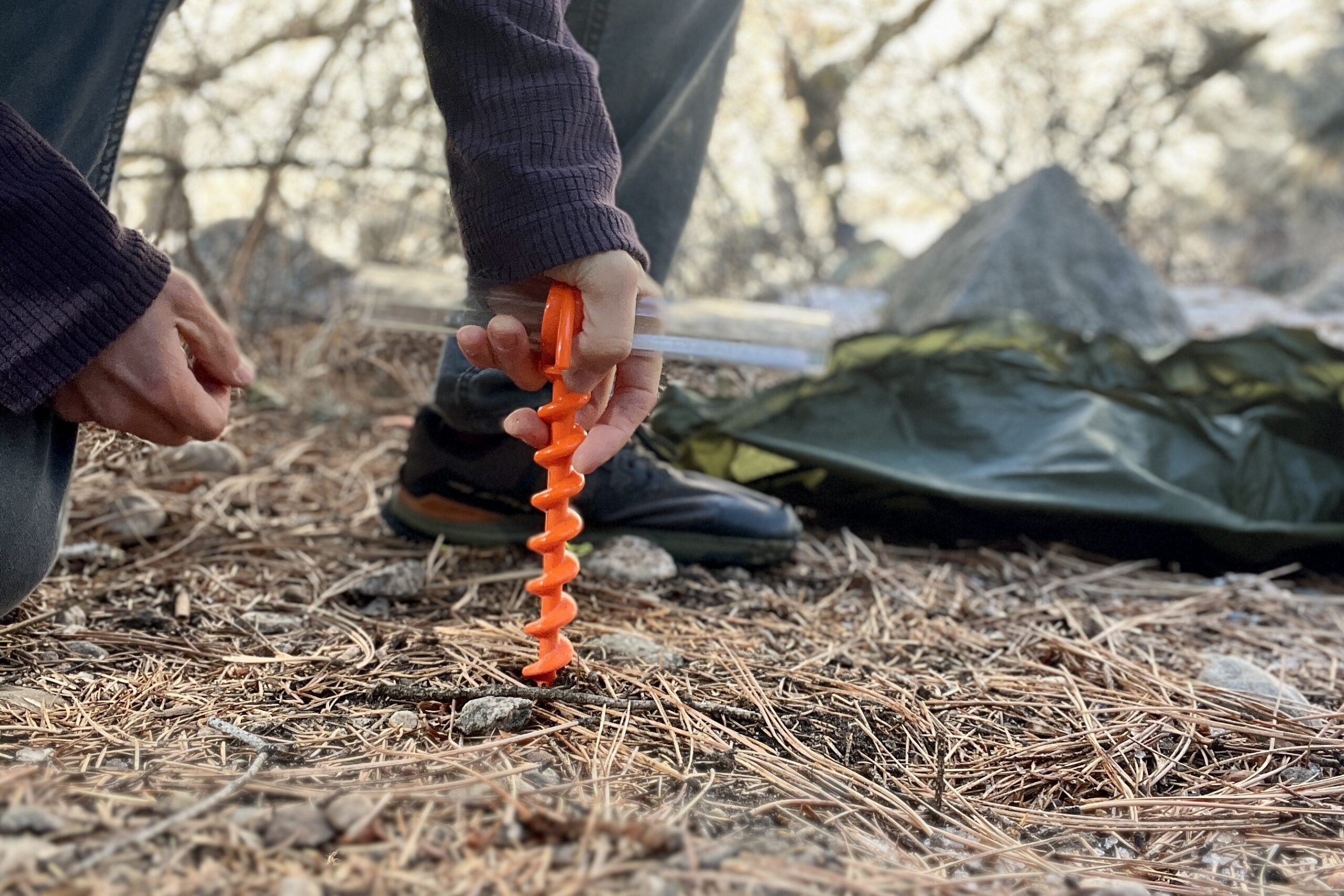 A hiker screws a corkscrew tent stake into piney soil with a tent in the background.