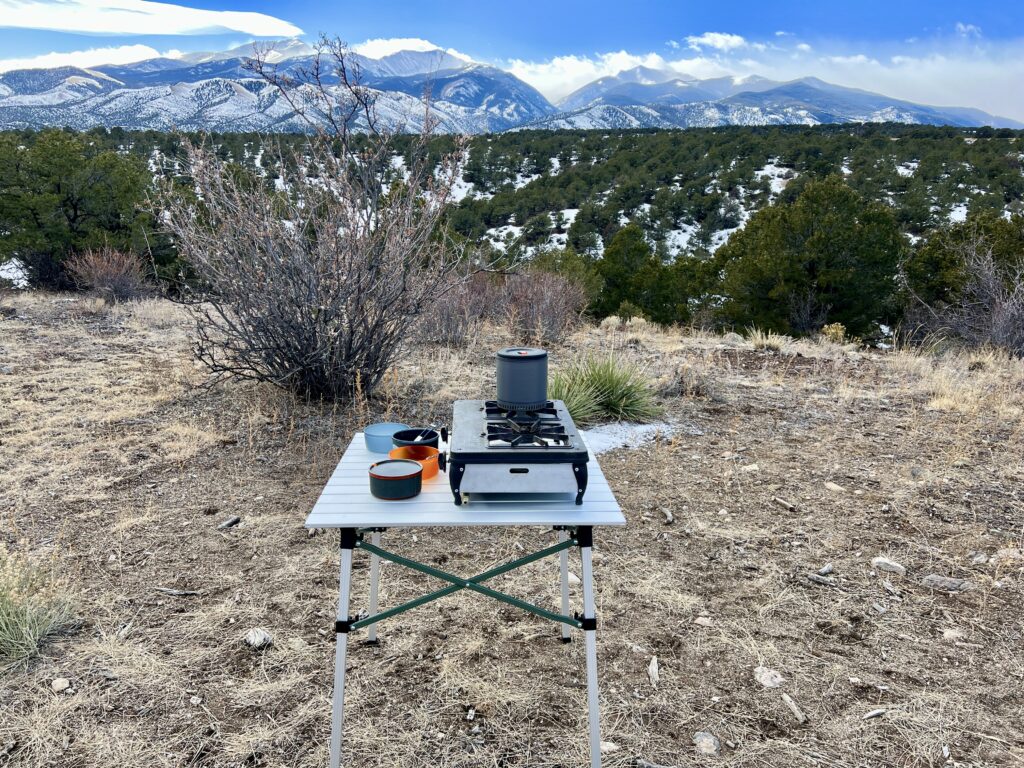 The REI Camp Roll Table stands outside with a mountain scene in the background. It has a two-burner stove and bowls sitting on its top.