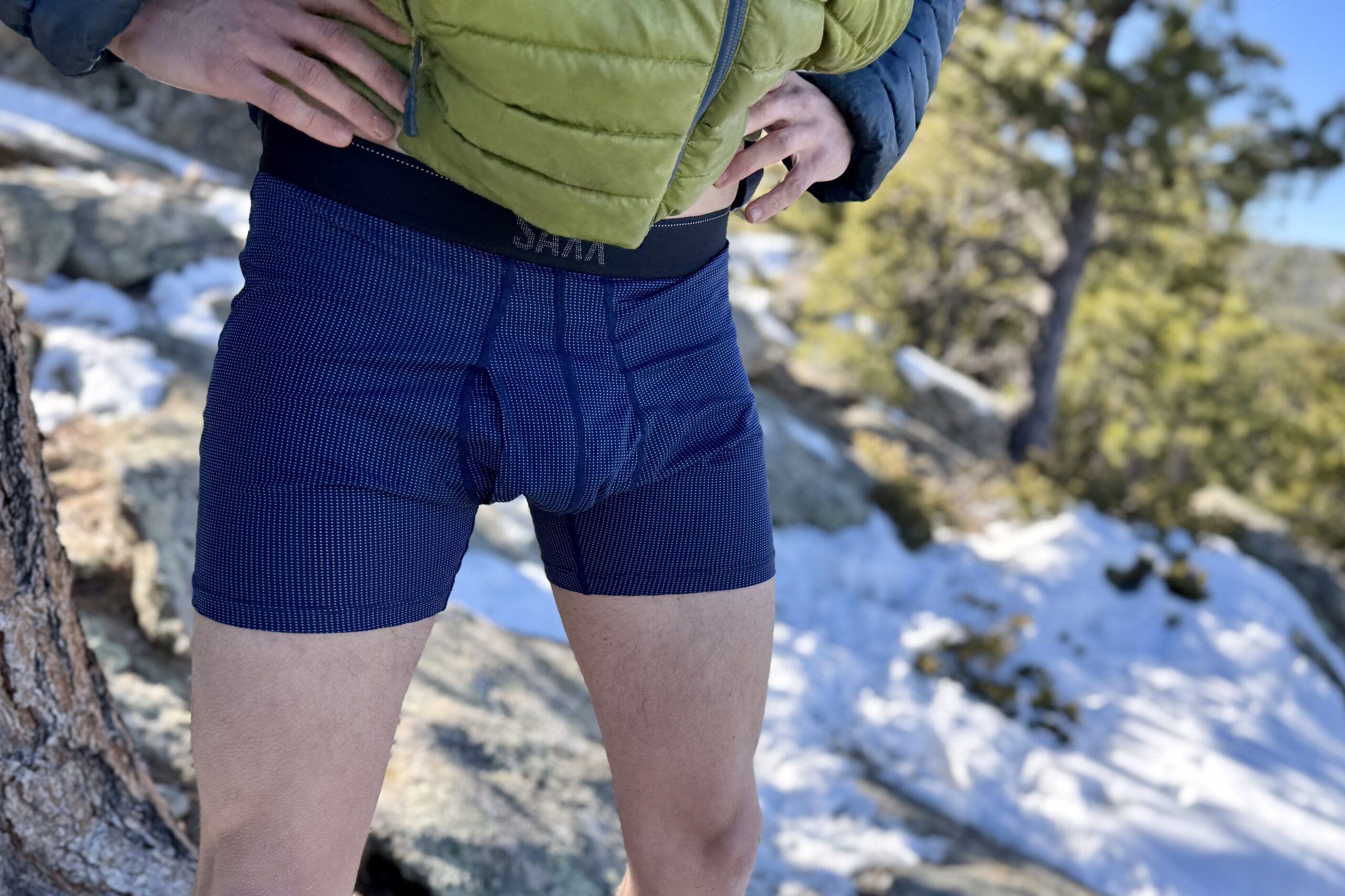A hiker stands in a snowy forest wearing nothing but his undies and a puffy jacket