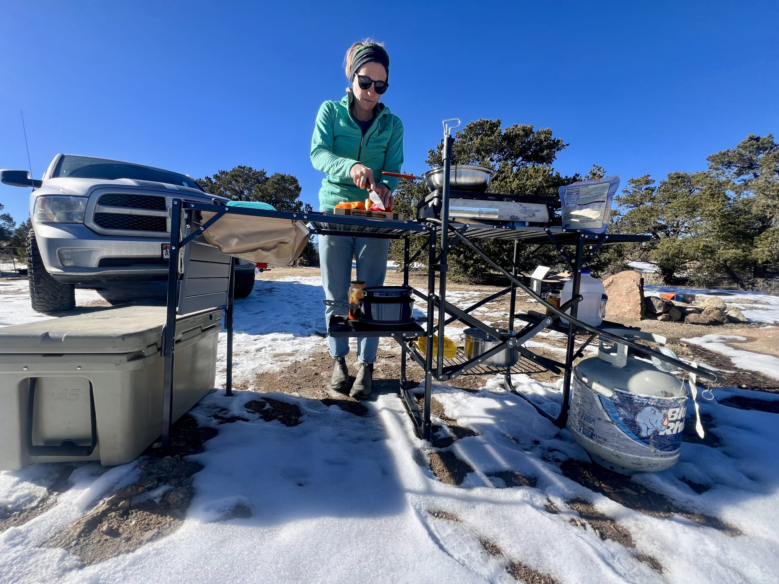 A person stands outside with a blue sky above them at the GCI Master Cook Station cutting produce. There is a cooler next to the station, snow on the ground, and a truck parked in the background.