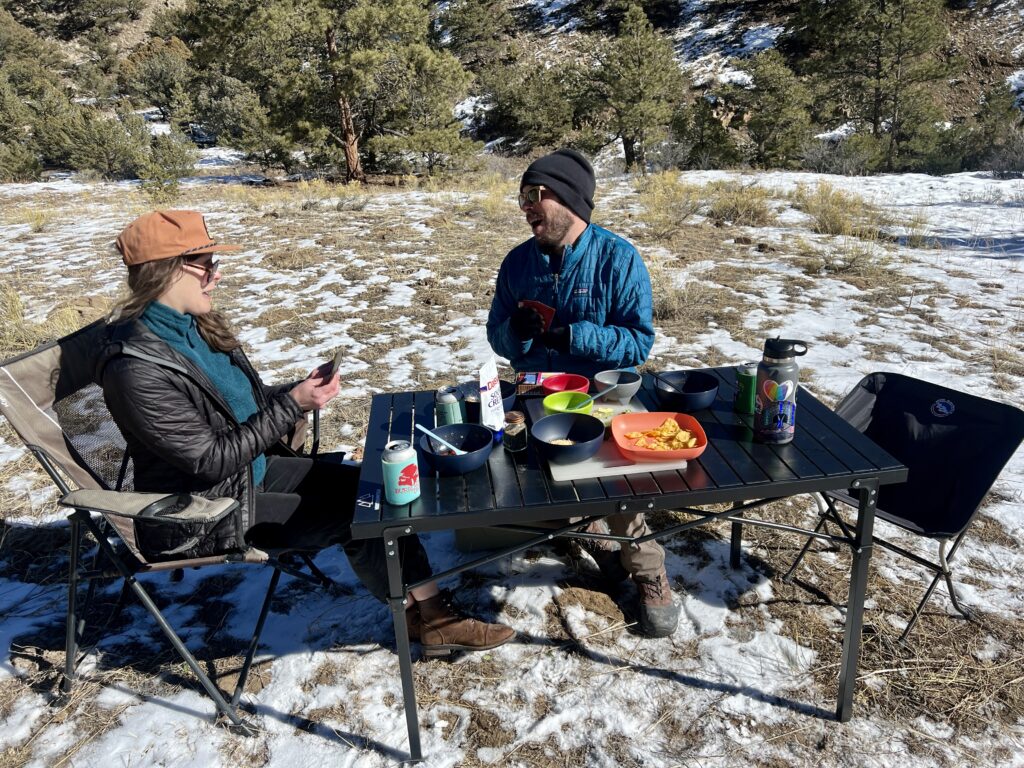 Two people sit and smile around the Mountain Summit Gear Heavy Duty Roll Top Table. They are wearing black and blue jackets and black and orange hats. They have playing cards in their hands. There are bowls, food, and drinks on the table.