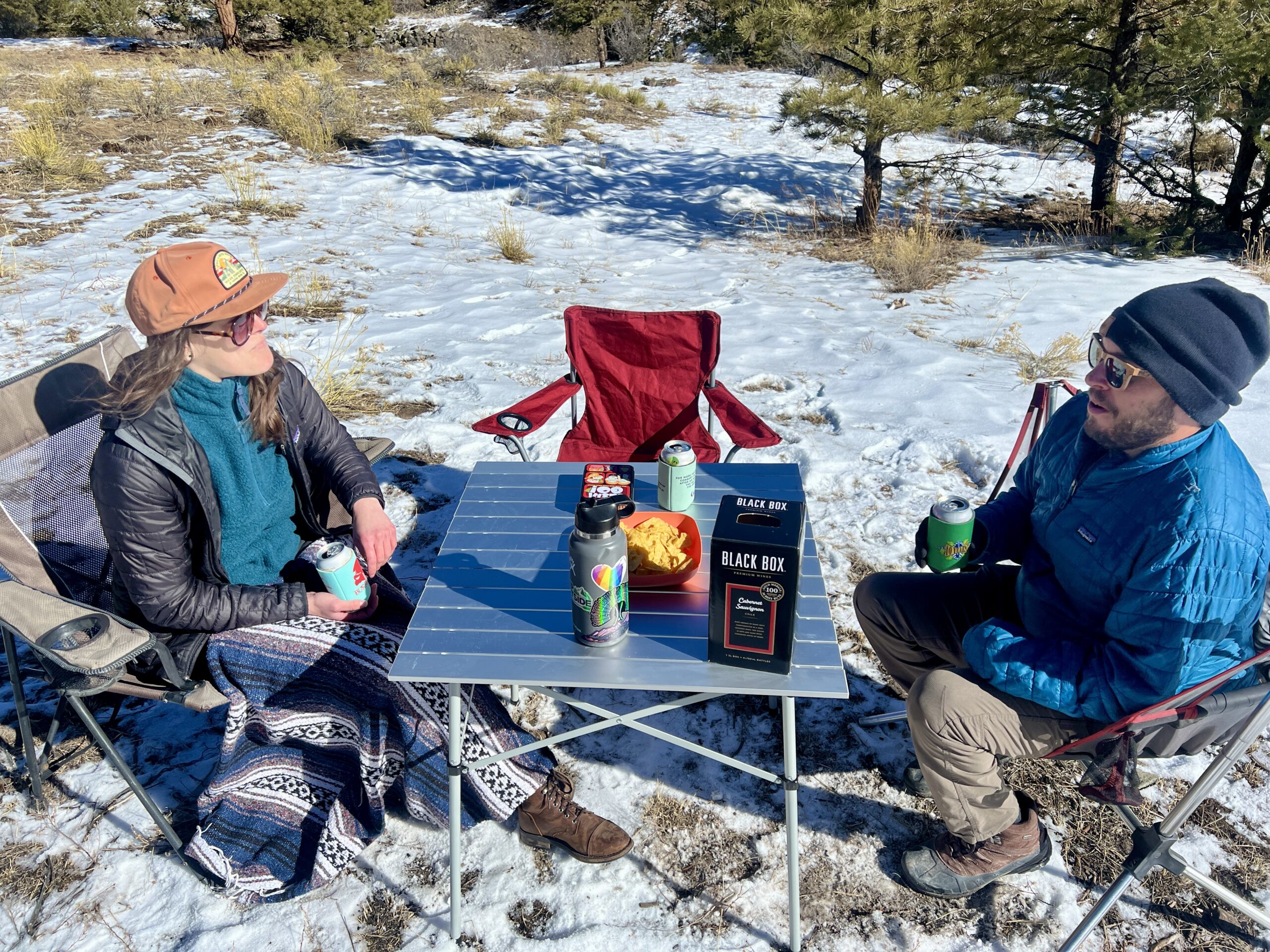 Two people sit around the Coleman Compact Roll-Top Table smiling. The background is snow and pine trees. The table has a box of wine, a water bottle, and chips.