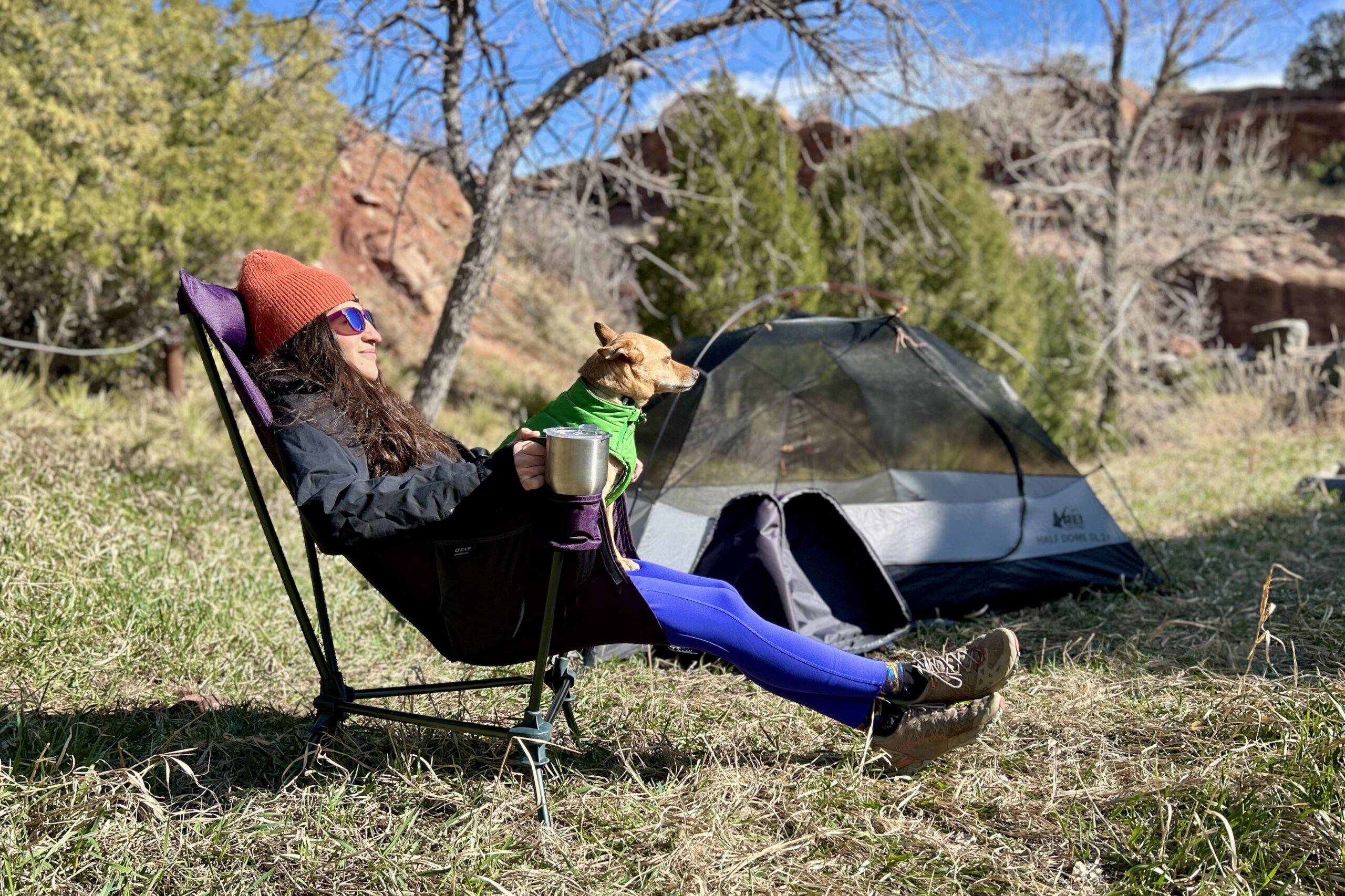 A woman leans back in a camping chair in a forested setting with her hand on a large mug and a small dog on her lap.