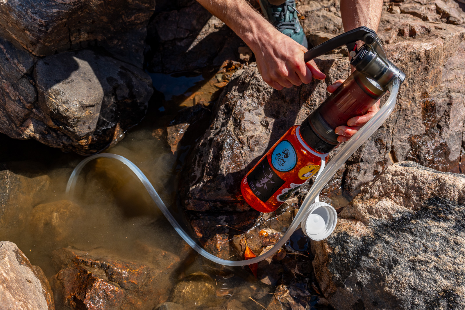 A hiker pumps with the msr guardian filter. the pre filter is submerged in murky water and a nalgene bottle is attached to the bottom of the filter.