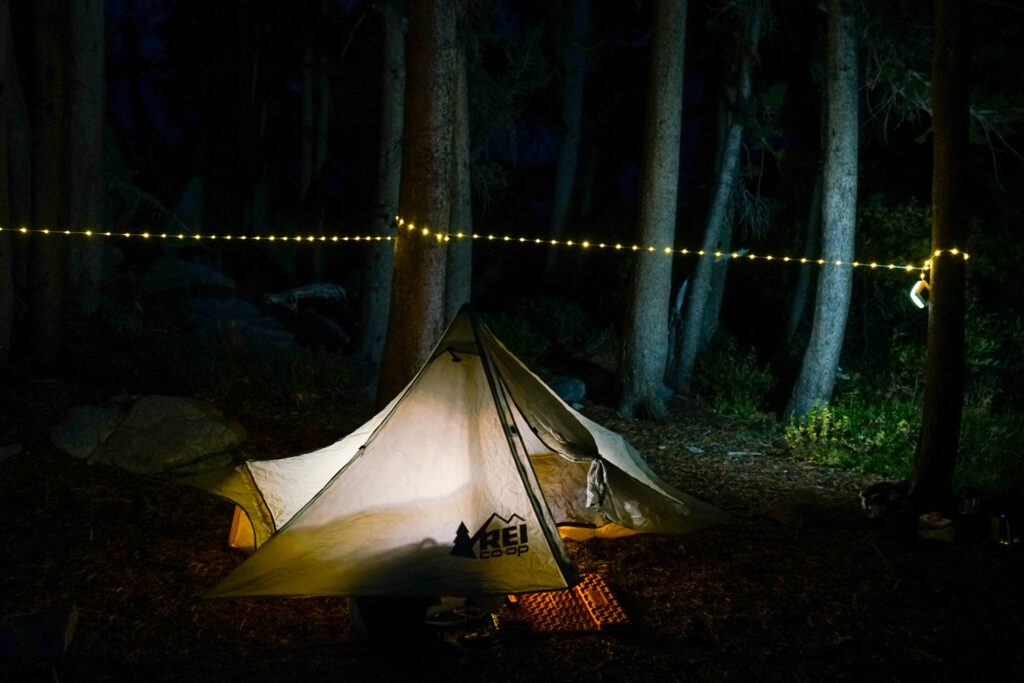 A glowing tent sits in the dark in a forest. The string light component of the LuminAid Solar String Light wraps around several trees behind the tent.