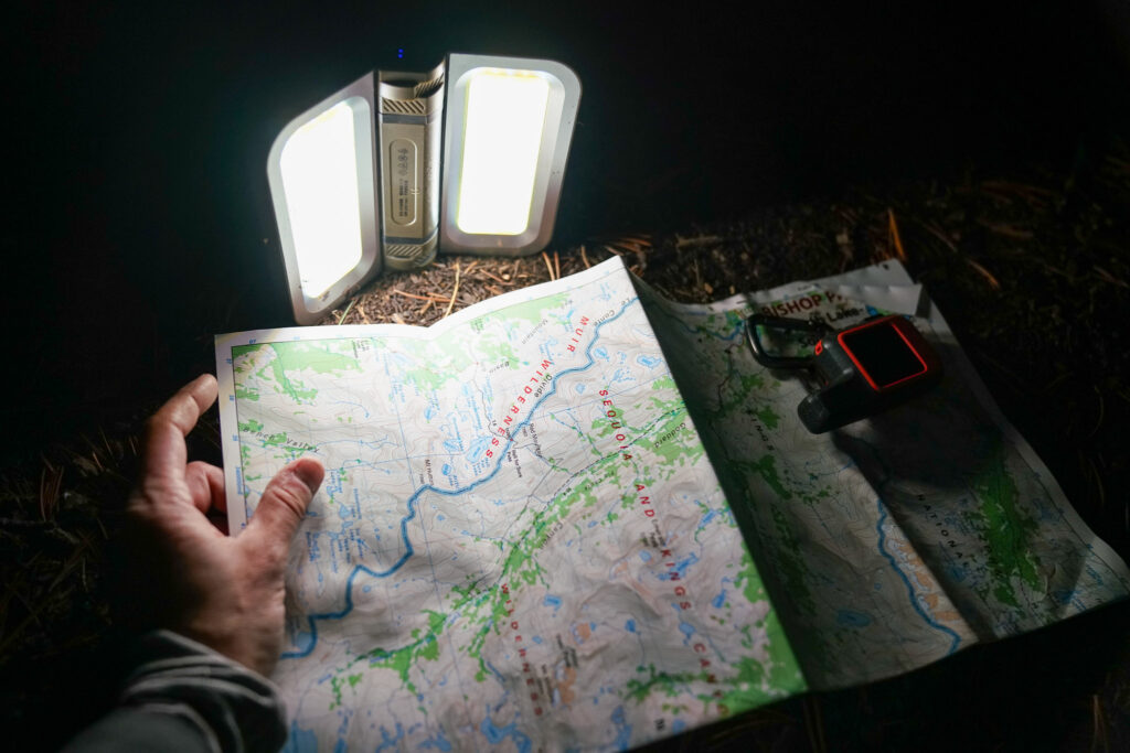 The LightUpCamp LED Solar Powered Foldable Camping Light sits on the ground, folded open and turned on to illuminate a map. A hand holds a corner of the map and a Garmin InReach device sits on the other corner.