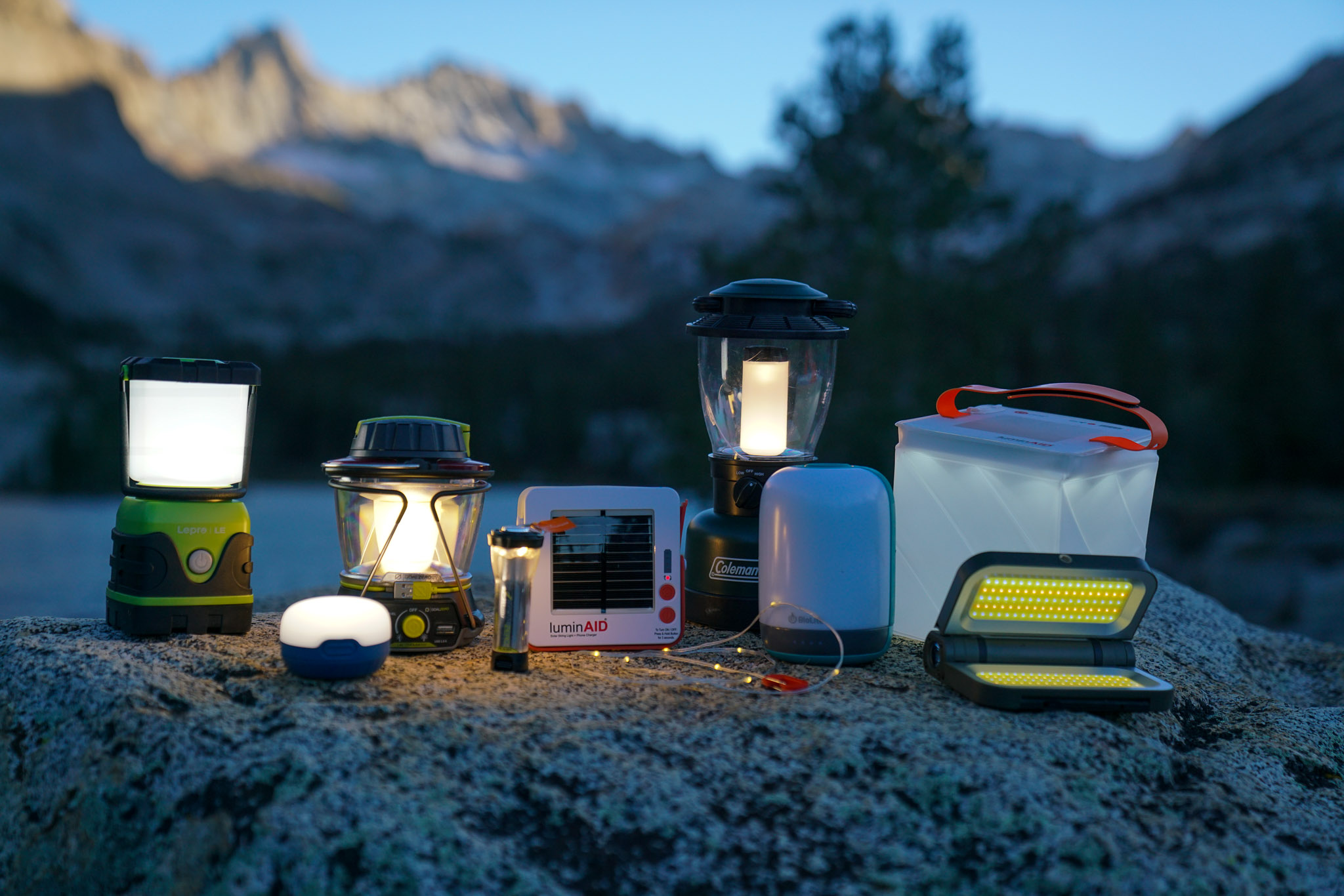 Nine lanterns- the LE 1000LM LED lantern, Black Diamond Moji, GoalZero Lighthouse 600, GoalZero Lighthouse Micro Flash, LuminAid String Light, Coleman Rugged Rechargeable, BioLite AlpenGlow 500, LuminAid Packlite Max 2-in-1, and LightUpCamp LED Solar Powered Foldable Camping Light- sit beside each other on a rock. The last remaining daylight illuminates the peaks of mountains in the background.