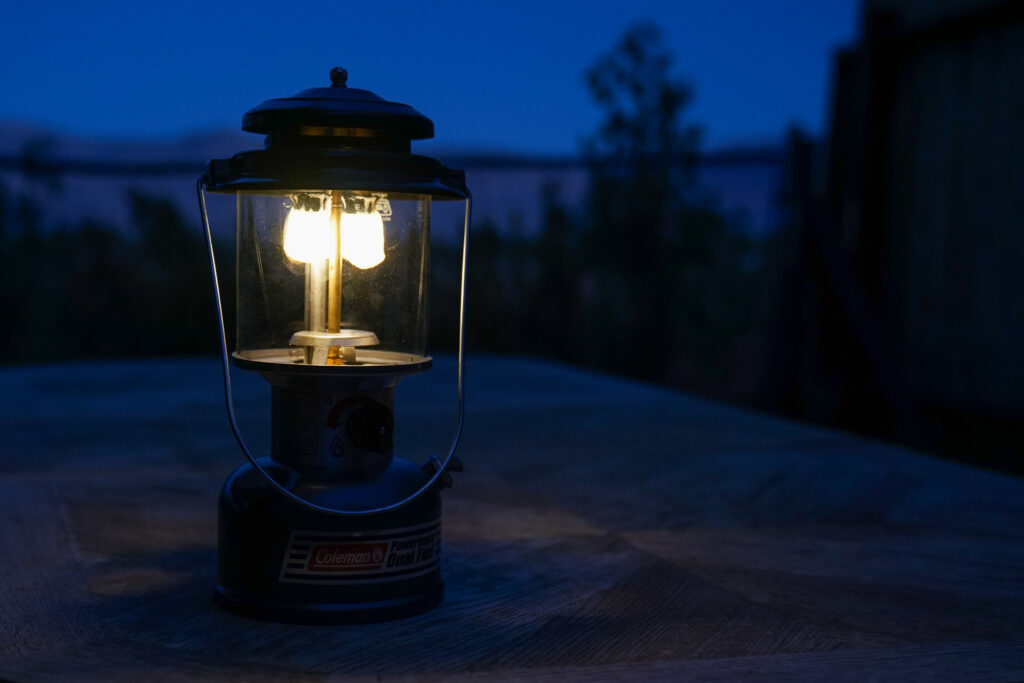 The Coleman Powerhouse Dual Fuel lantern sits in the twilight on a table. The lantern's mantles glow softly.