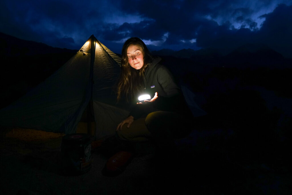 A person crouches on the ground holding the Black Diamond Moji lantern in their hand. In the background is an illuminated tent and cloudy mountaintops.