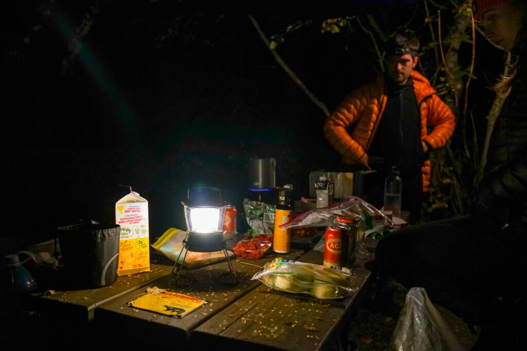 The GoalZero Lighthouse 600 lantern sits on a picnic table with food and drinks around it. A person stands at one end of the table watching a pot on a stove.
