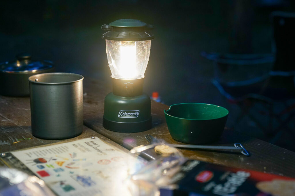 The Coleman Rugged Rechargeable lantern sits on a picnic table. The lantern is turned on, providing a warm light, with a bowl and pot sitting beside it.